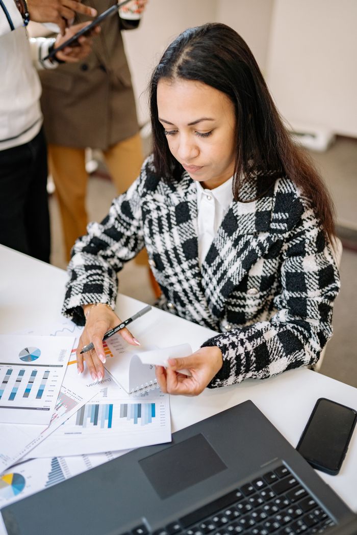 Focused businesswoman reviewing financial documents at her desk with charts and laptop.